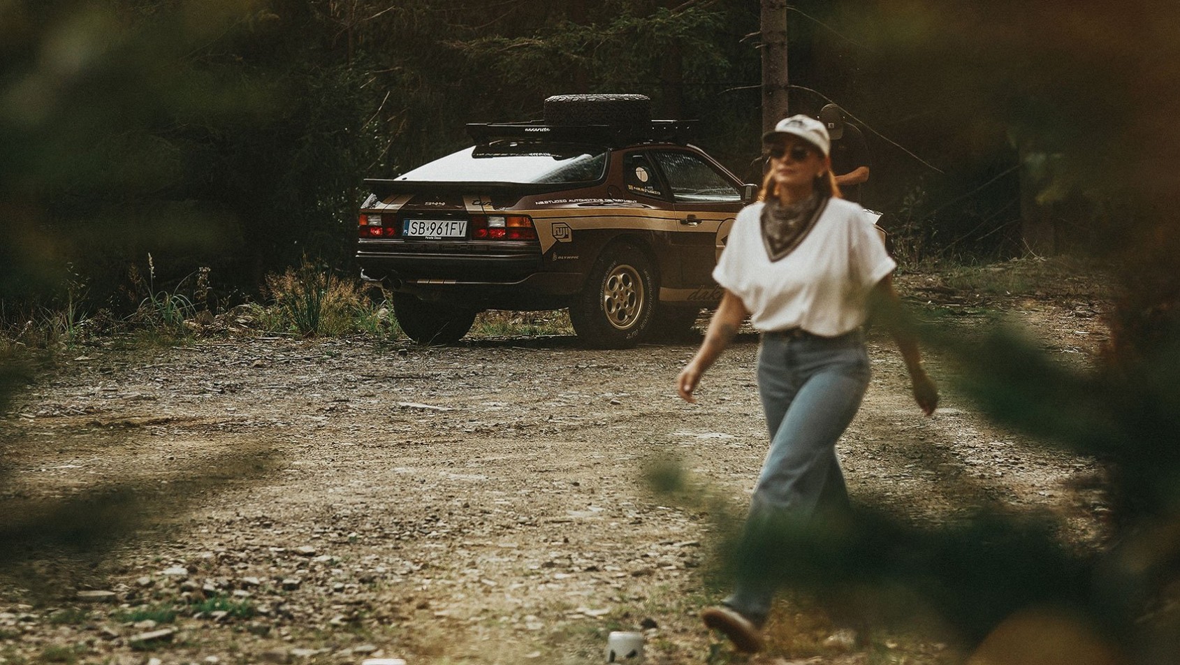 Porsche 944 Dakarista zaparkowane w lesie, kobieta w kapeluszu odchodzi w kadrze