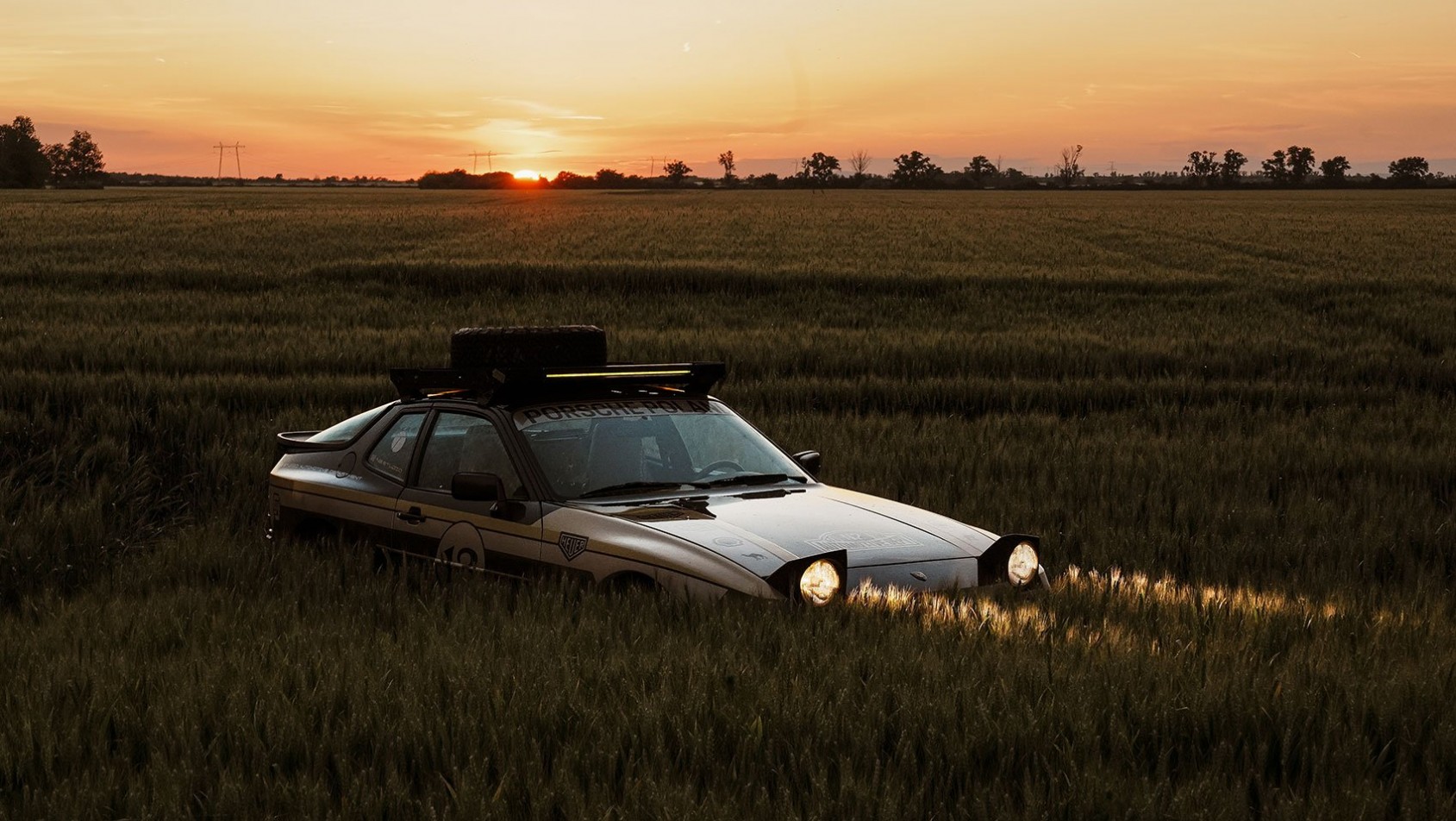 Porsche 944 Dakarista z włączonymi światłami na tle zachodzącego słońca i pól zboża