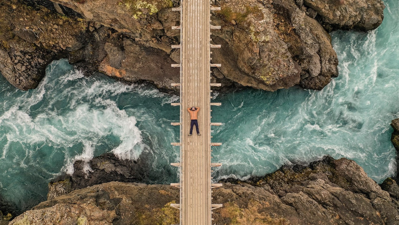 Mężczyzna leżący na drewnianym moście nad rwącą, błękitną rzeką – zdjęcie z drona