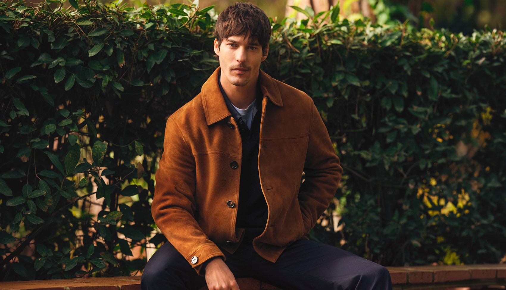 Mężczyzna w brązowej kurtce skórzanej OCHNIK siedzący na ławce wśród zieleni Brązowa kurtka skórzana OCHNIK – casualowa elegancja