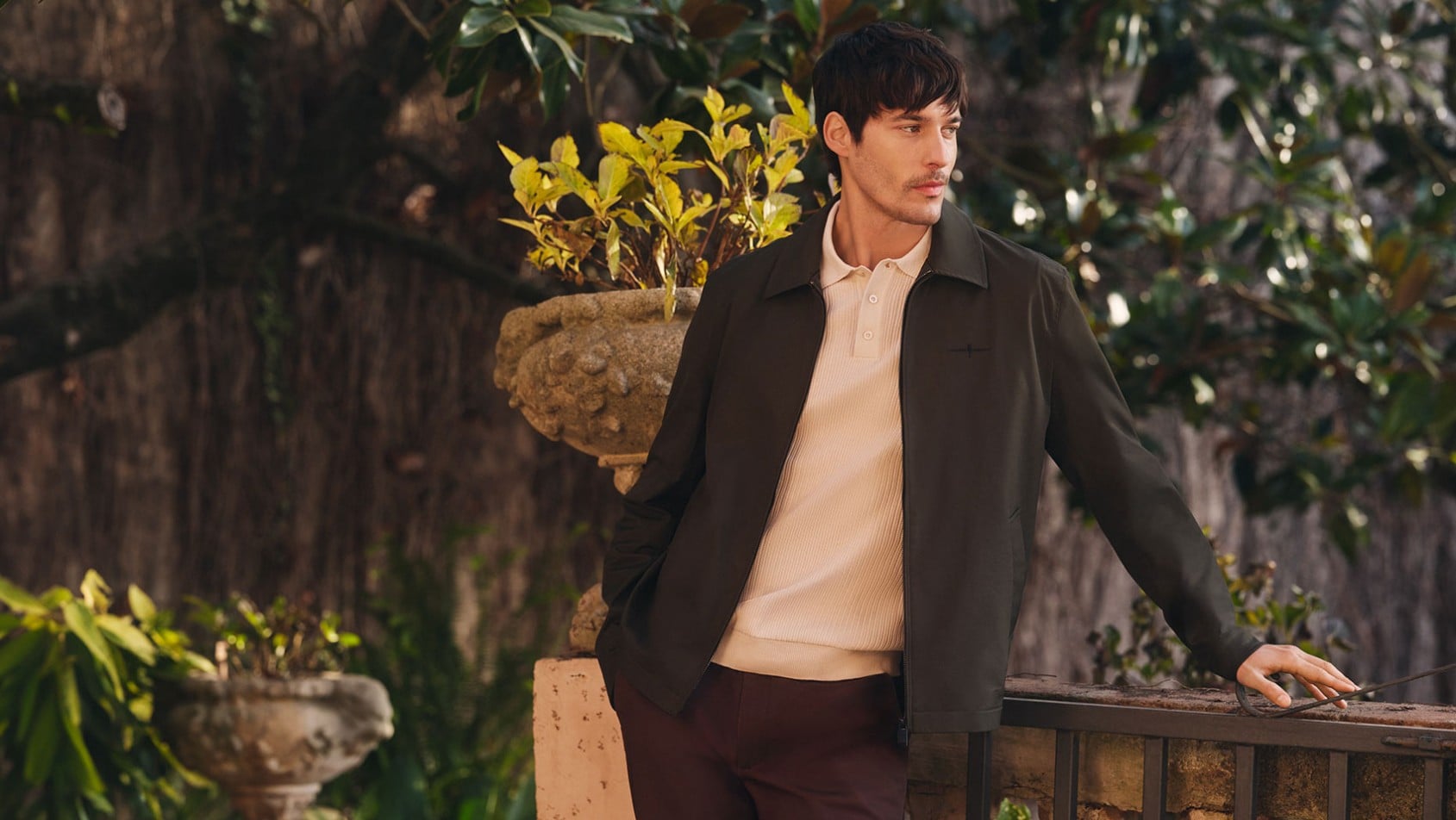 Mężczyzna w zielonej kurtce OCHNIK i beżowym polo oparty o balustradę w ogrodzie