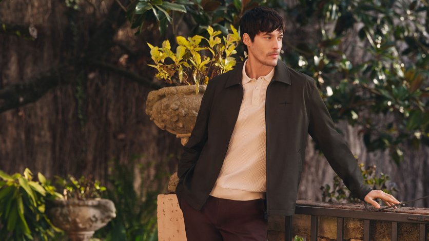 Mężczyzna w zielonej kurtce OCHNIK i beżowym polo oparty o balustradę w ogrodzie