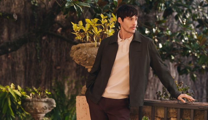 Mężczyzna w zielonej kurtce OCHNIK i beżowym polo oparty o balustradę w ogrodzie